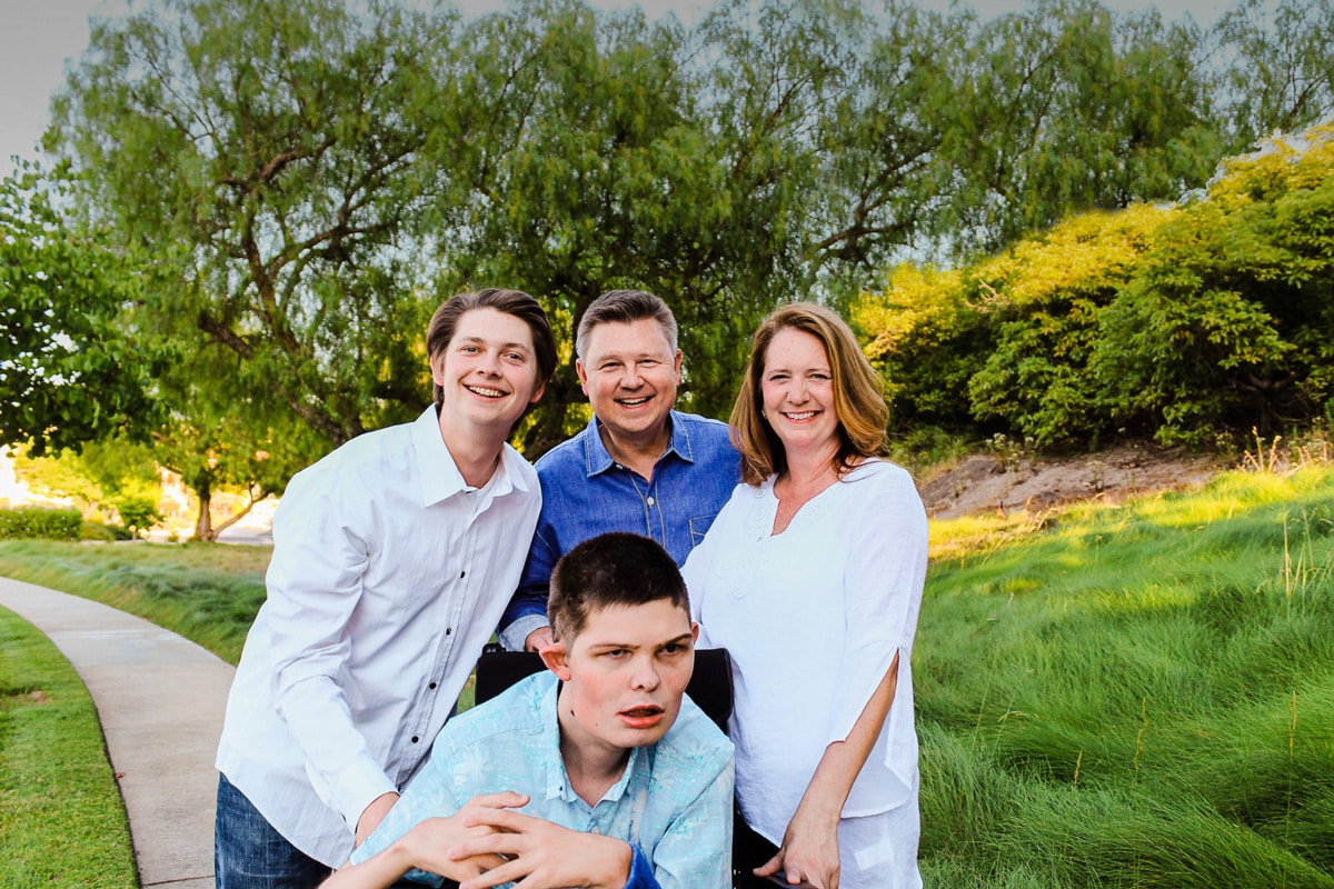 A family portrait of Steve Bundy and his family. His wife, Melissa is to his right, his younger son, Jaron is to his left and his son Caleb is in the front of the three in his wheelchair. They're all smiling at the camera with lush trees and grass in the background.
